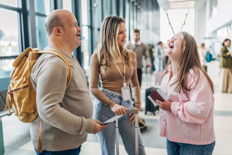 Two individuals with Down syndrome laugh and talk with their caregiver at the airport. They hold travel documents and seem excited about their trip. People are nearby.