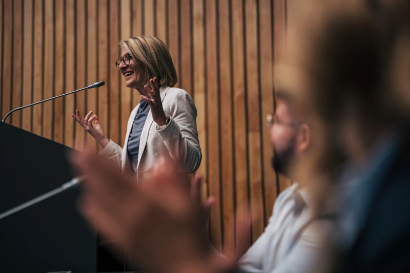 Mature adult woman standing at a podium delivering a speech to an audience, actively engaging and gesturing, receiving applause from attendees at a corporate event or seminar
