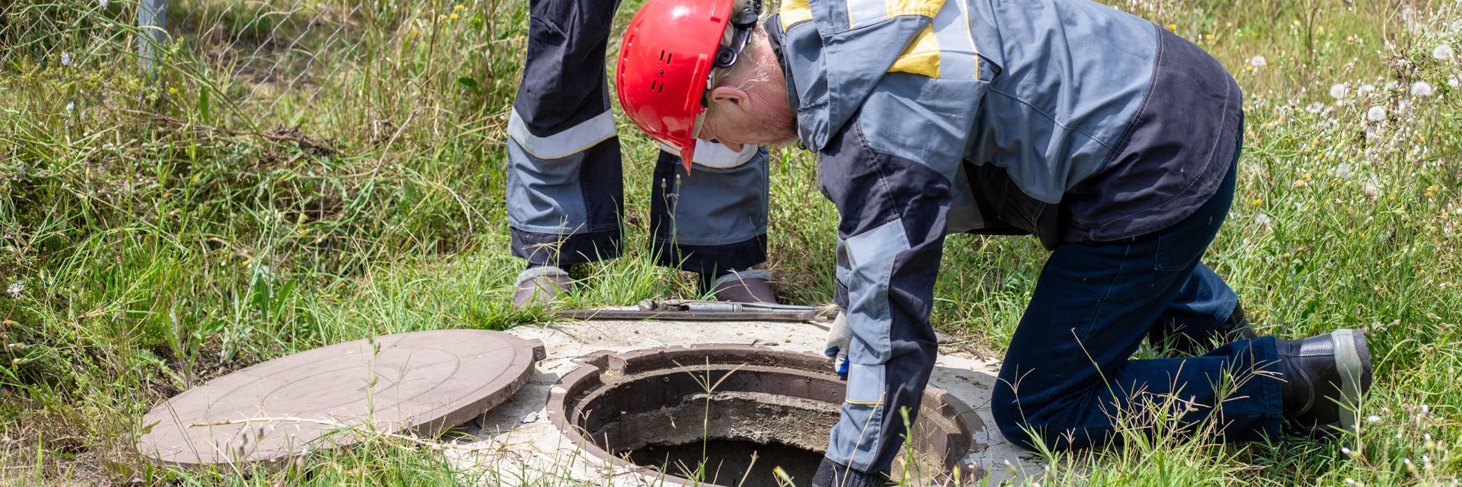 Two workers in hard hats checking an open manhole from outside before entry