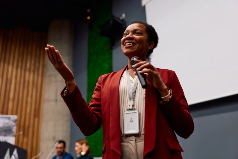 Confident woman speaking at business conference, holding microphone and gesturing, engaging audience with presentation