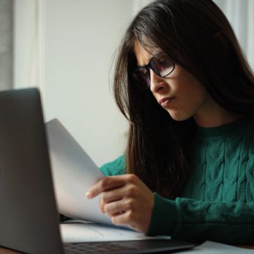 Woman reading a document with a laptop open.
