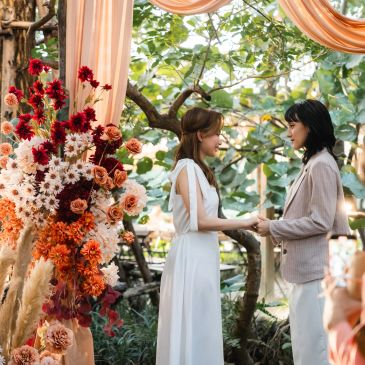 Pareja sosteniéndose las manos rodeada de flores en una ceremonia al aire libre.