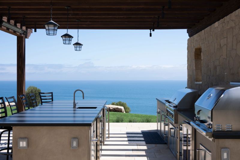 A high-end outdoor kitchen setup featuring stainless steel grills and a spacious island with a sink, overlooking a vast blue ocean horizon