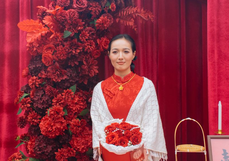 Gentle Asian bride dressed in traditional red cheongsam and white knitted shawl holds red rose gift box in front of red floral arrangement and velvet curtain, recording warm precious moments of classic traditional Chinese wedding cultural lifestyle.