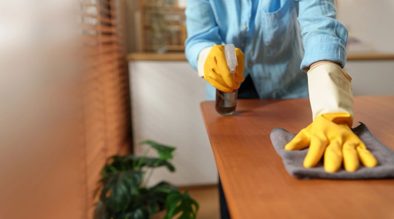 Woman wipes a wooden table with a microfiber cleaning cloth while using disinfectant spray, promoting hygiene, cleanliness, and efficient household care in a modern home.