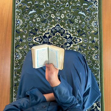Person reading a Quran, learning about Islam on a patterned green carpet.