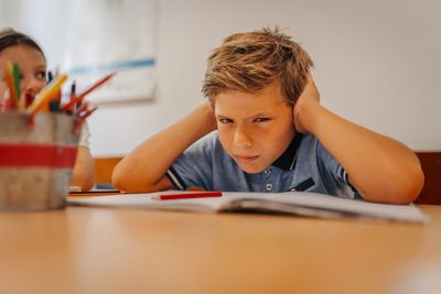 Frustrated boy covering ears at school desk.