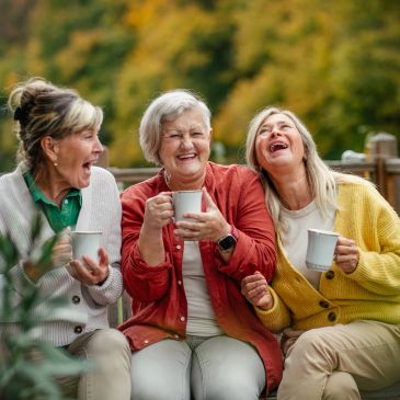Three older women enjoying coffee and laughing outdoors.