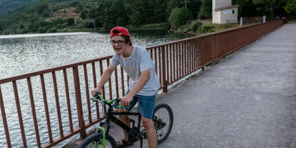 Boy riding bike near water and fence.