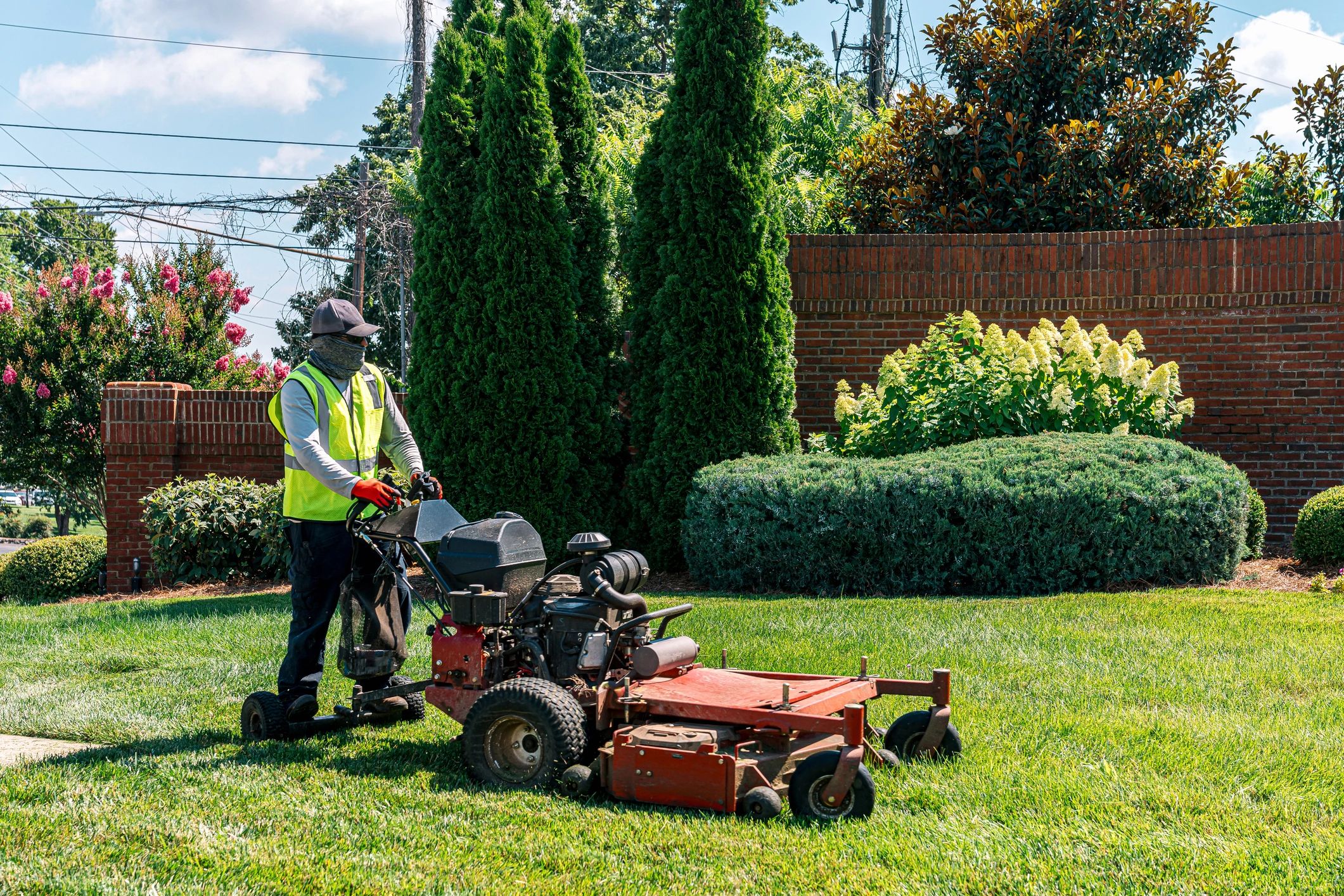 Worker mowing grass near brick wall and trees.