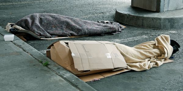 Two homeless people sleeping outdoors under blankets and cardboard.