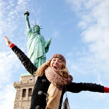 A lady poses smiling in front of the Statue of Liberty on a cold, clear day with blue skies.

