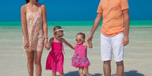 Family of four holding hands and walking on a sunny beach with turquoise water.