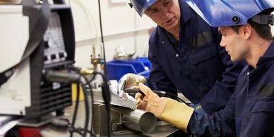 Two welders in protective gear working together on a metal pipe.
