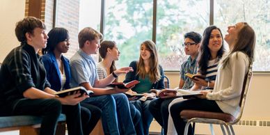 A diverse group of young adults holding books, engaged in an animated discussion indoors.