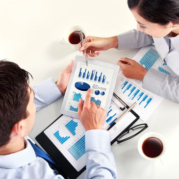 Two professionals analyzing blue bar and pie charts on a tablet and papers with coffee cups nearby.