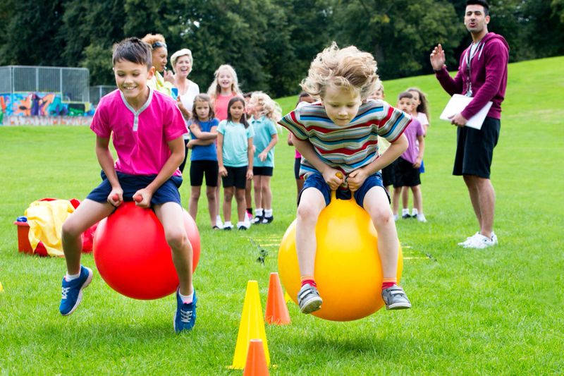 Little boys sitting on  space hoppers racing,  classmates stand in the background watching during a sports lesson.