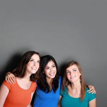 Three women smiling together against a gray background.