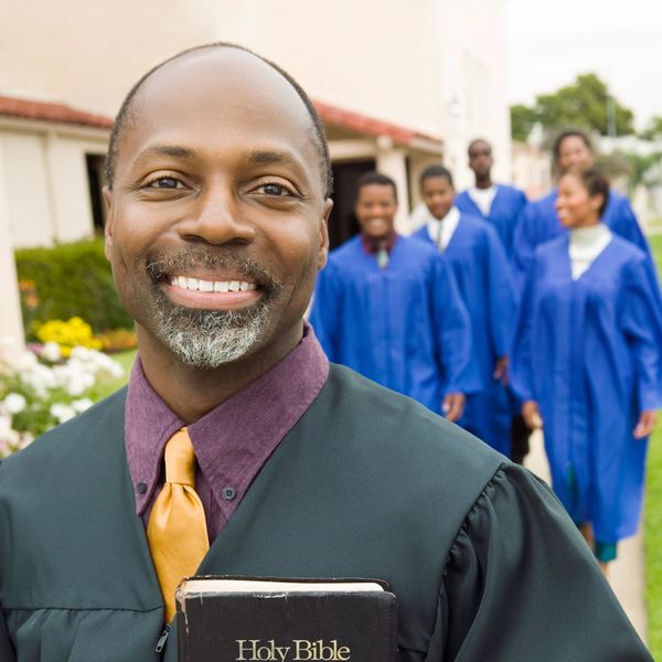 Smiling man in a robe holding a Bible with graduates in blue behind.