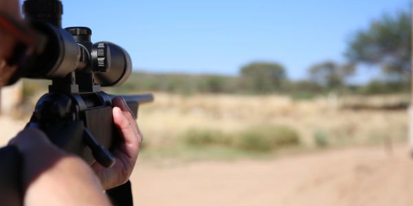 Person aiming a scoped rifle outdoors.