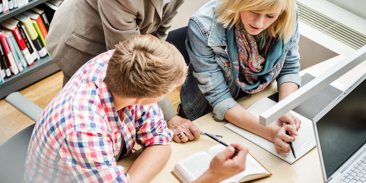 Three people studying together at a library desk with a computer and open book.
