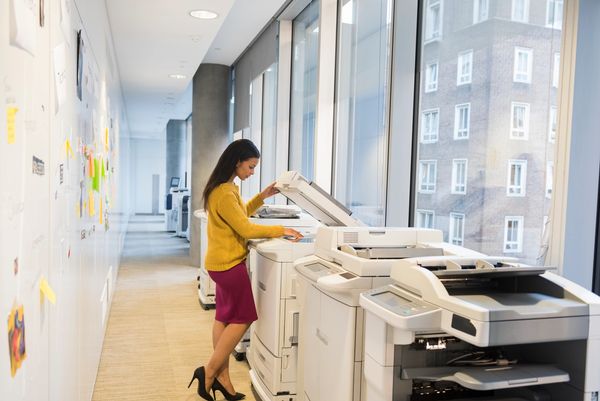 Woman using a large multi-function office copier/printer in a hallway with large windows.