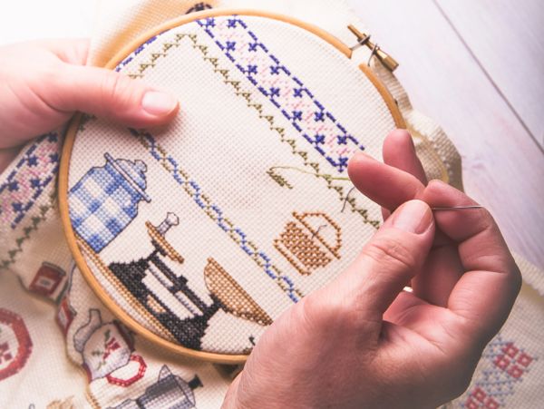Hands working on a detailed cross-stitch embroidery with kitchen-themed designs.