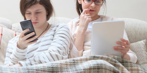 Two women sitting on a couch, one using a smartphone and the other a tablet.