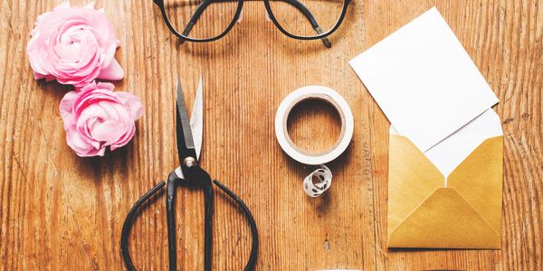 Crafting supplies including pink flowers, scissors, tape, glasses, and an envelope on a wooden table.