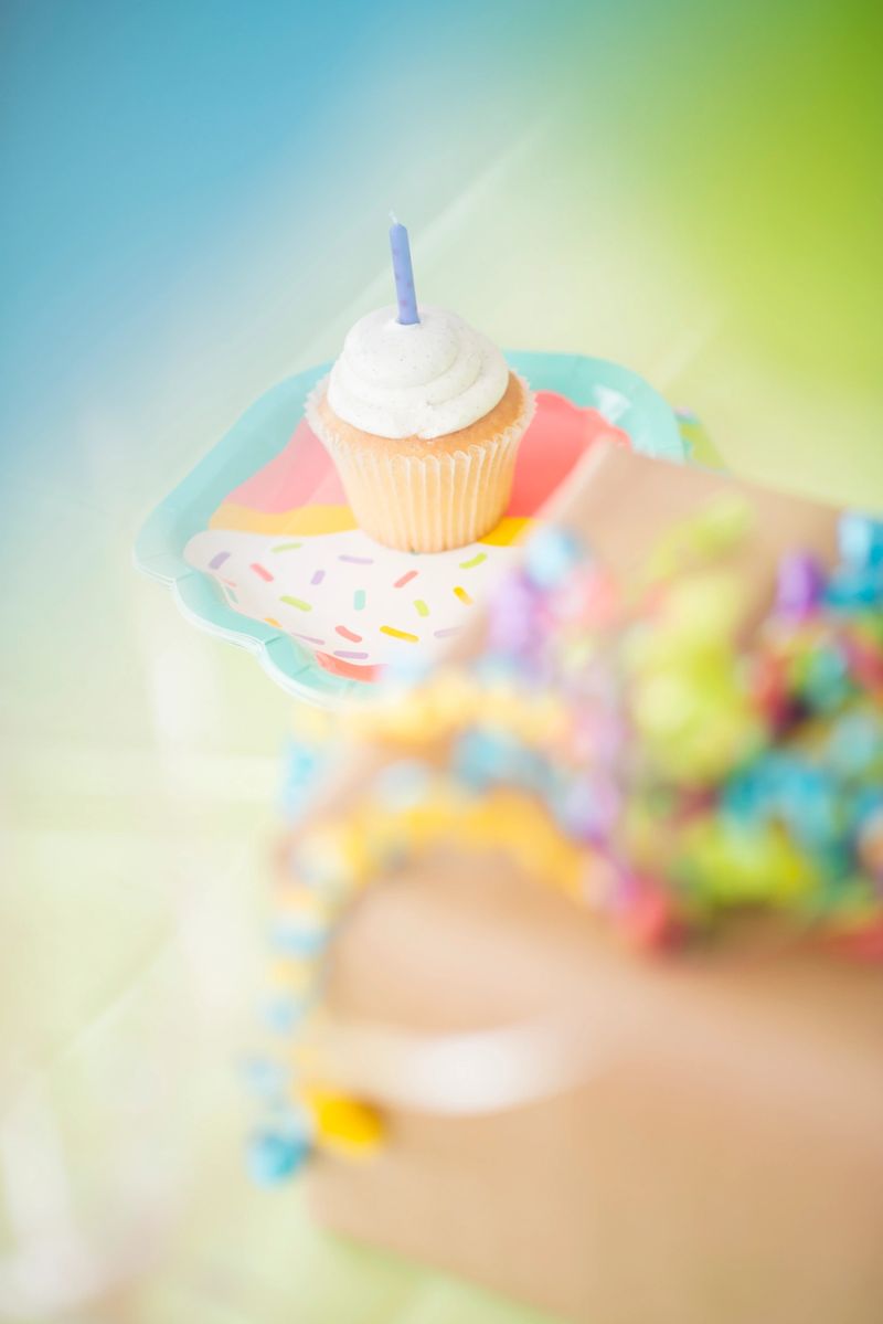 Single Vanilla bean cupcake with birthday candle on a cupcake plate. Birthday gifts and balloons out of focus in foreground.