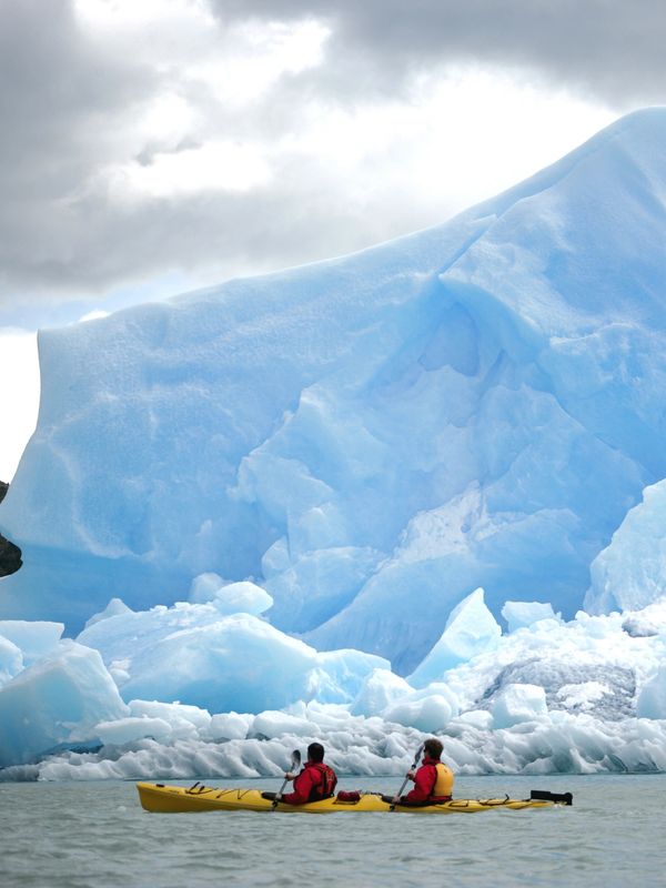 Two people kayaking near a massive blue iceberg under cloudy skies.