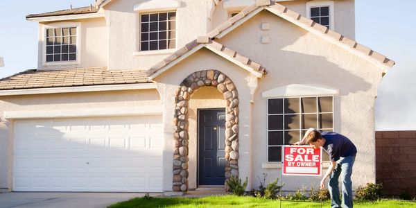 Person placing a 'For Sale by Owner' sign in front of a suburban house.