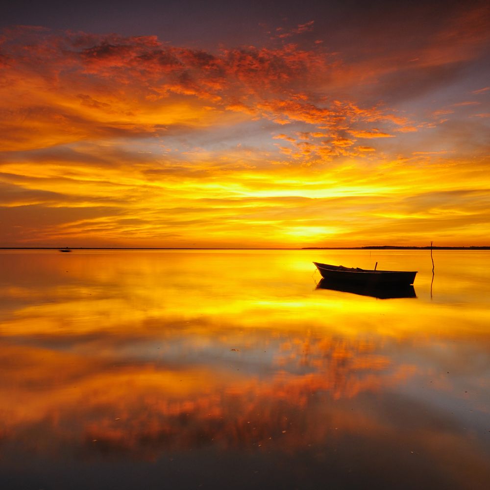 A lone boat floats on calm water under a vibrant orange sunset sky.