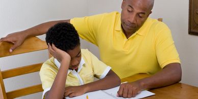 Father helps son with homework while boy looks frustrated.