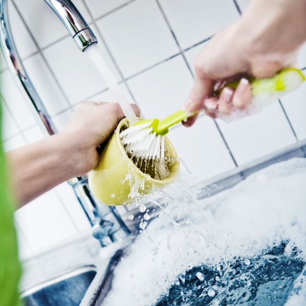 Person washing a yellow cup with a brush under running water in a kitchen sink.