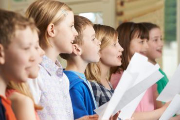 Children singing together while holding sheet music in a classroom.