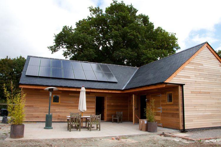 Modern wooden house with solar panels on the roof and outdoor seating area.
