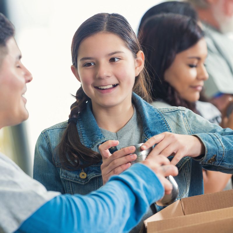 Preteen Caucasian Hispanic mixed race girl is volunteering in community food bank for charity. She is sorting donated food into cardboard boxes so they can be distributed for charity. Diverse volunteers are sorting grocery donations behind her in assebly line. 
