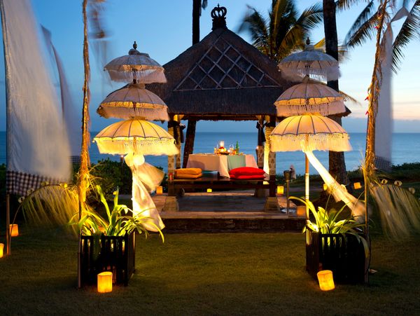 Romantic outdoor dining setup by the ocean at dusk with decorative umbrellas and soft lighting.