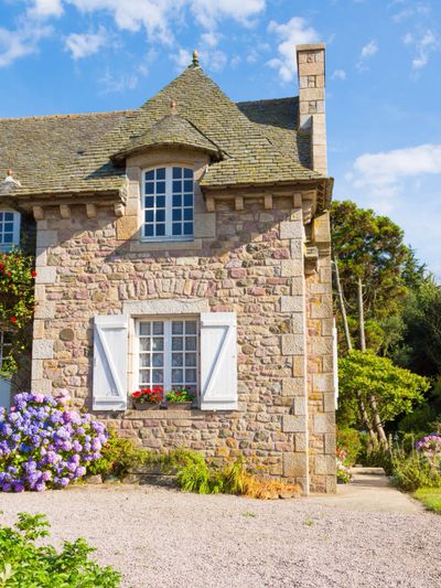 Charming stone cottage with white shutters and blooming flowers under a blue sky.