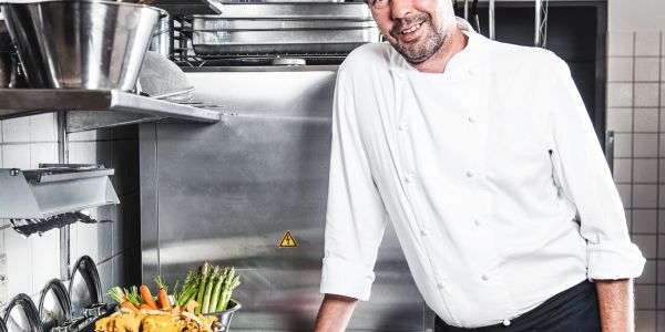 Chef in white uniform standing in kitchen with fresh vegetables.