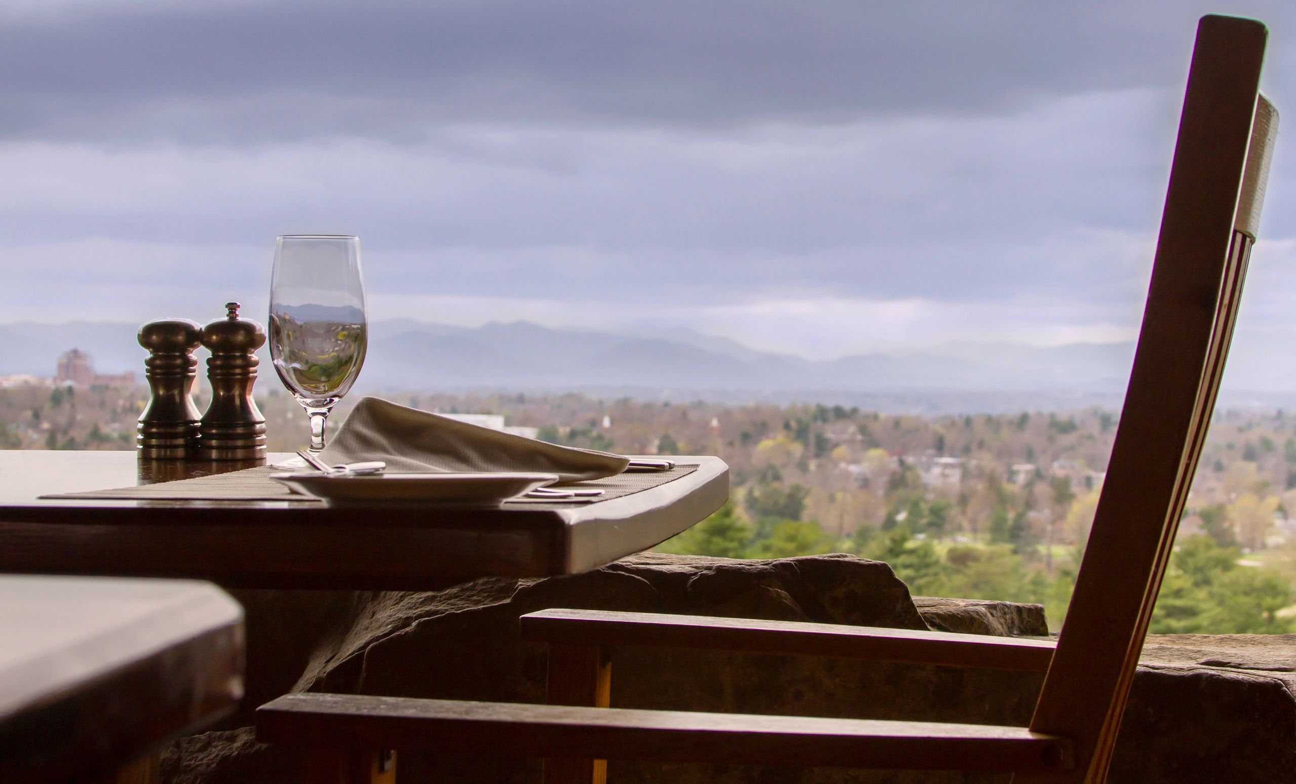 A view mountains in the distance through a wine glass that is set up with a dinner set up on a table. Also on the table are salt and pepper shakers, a cloth napkin, small plate and silverware. A wooden chair on the right.