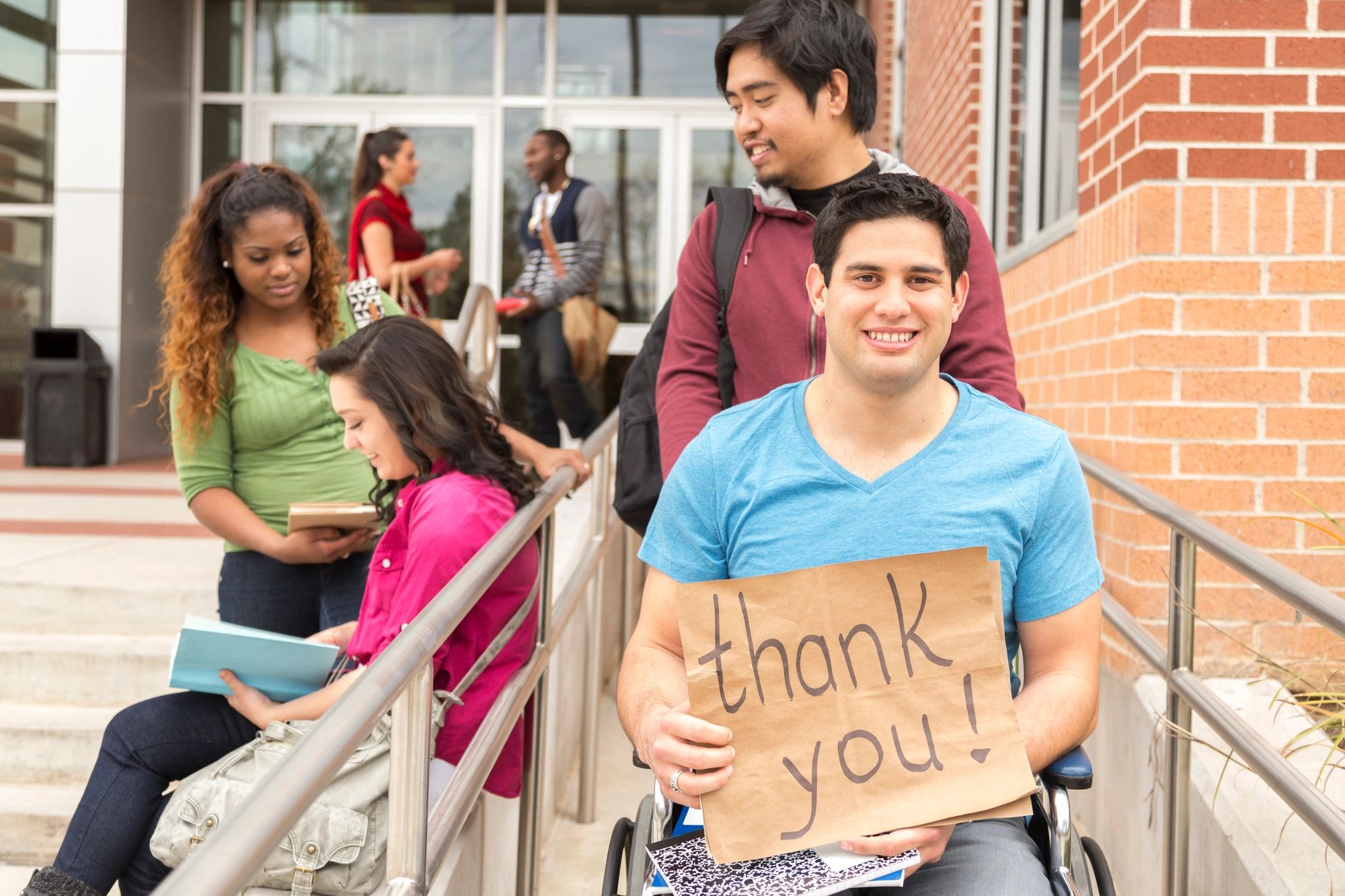 A young man sitting in a wheelchair is holding a paper sign that says, "Thank you." He is being push