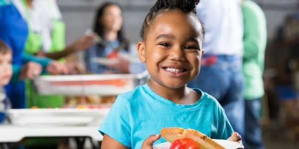 Smiling child holding a plate with bread and an apple in a busy food line.