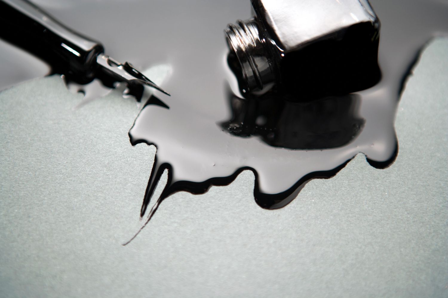 A fountain pen next to spilled black ink on a gray surface.