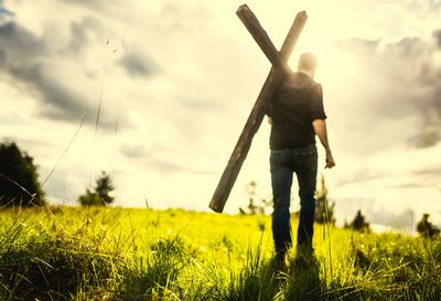 Man carrying a wooden cross on a sunlit grassy hill under a cloudy sky.