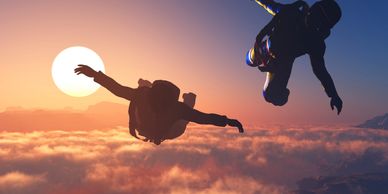 Two skydivers silhouetted against a glowing sunset above clouds.