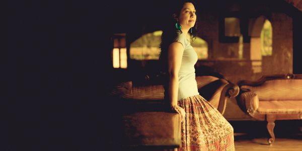 Woman sitting on a couch in warm, dim lighting, wearing a patterned skirt.
