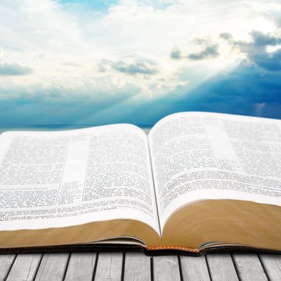 Open Bible on wooden surface with sun rays breaking through clouds in background.