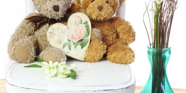 Two teddy bears with a heart and flowers on a white chair beside a green vase.